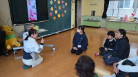 全員が床に座って教員の説明を聞いている。電子黒板の画面上には３年生児童が写っている。