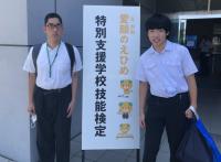 技能検定の看板の前に立つ２人の男子生徒。