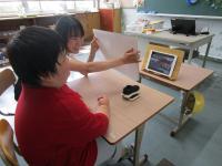 クイズを出している場面。みどり小学校の児童の映っているiPadに向けて教師がクイズをフリップを出し、児童が問題を言っている。